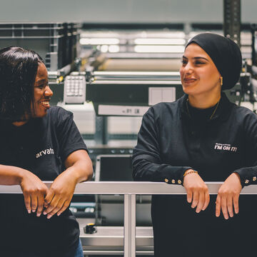 Zwei Mitarbeiterinnen lehnen locker an  einem Geländer und lachen miteinander