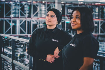 Zwei Mitarbeiterinnen in Arvato Kleidung lehnen lässig an einem Geländer und lassen ihren Blick in die Ferne schweifen