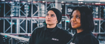 Zwei Mitarbeiterinnen in Arvato Kleidung lehnen lässig an einem Geländer und lassen ihren Blick in die Ferne schweifen