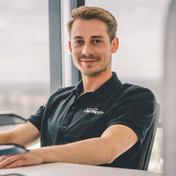 An employee wearing an Arvato polo shirt sits at a desk and looks into the camera